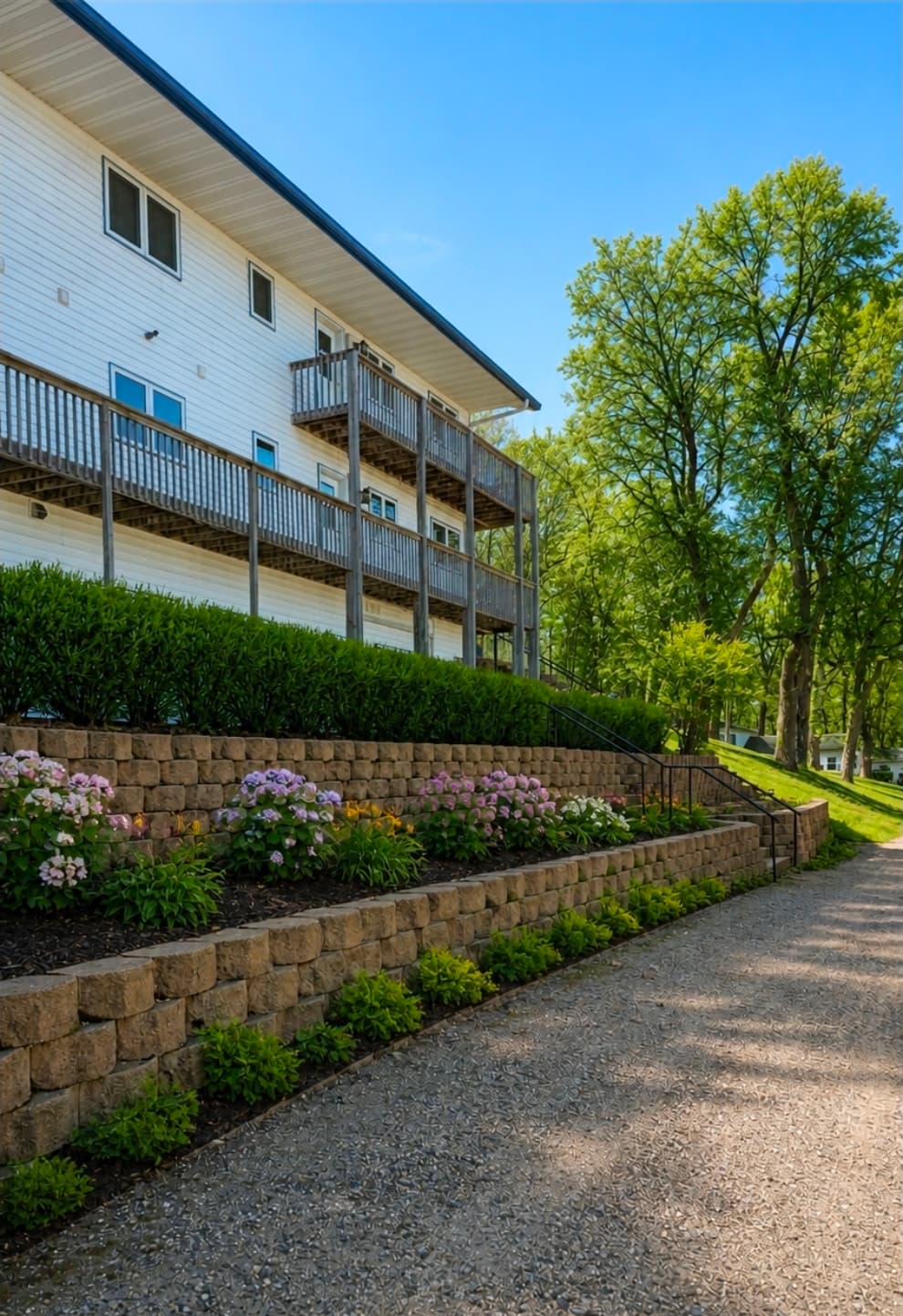 A multi-level white building with balconies surrounded by green shrubs and flower beds along a gravel pathway.