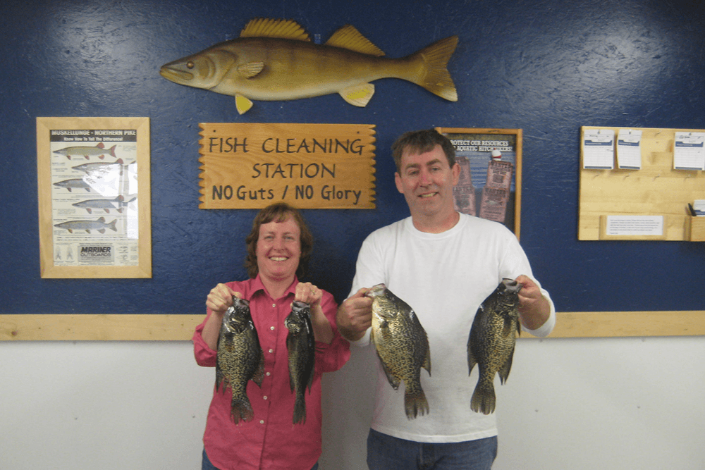 Two people holding fish in the fish cleaning area, with a sign behind them that reads Fish cleaning station no guts no glory