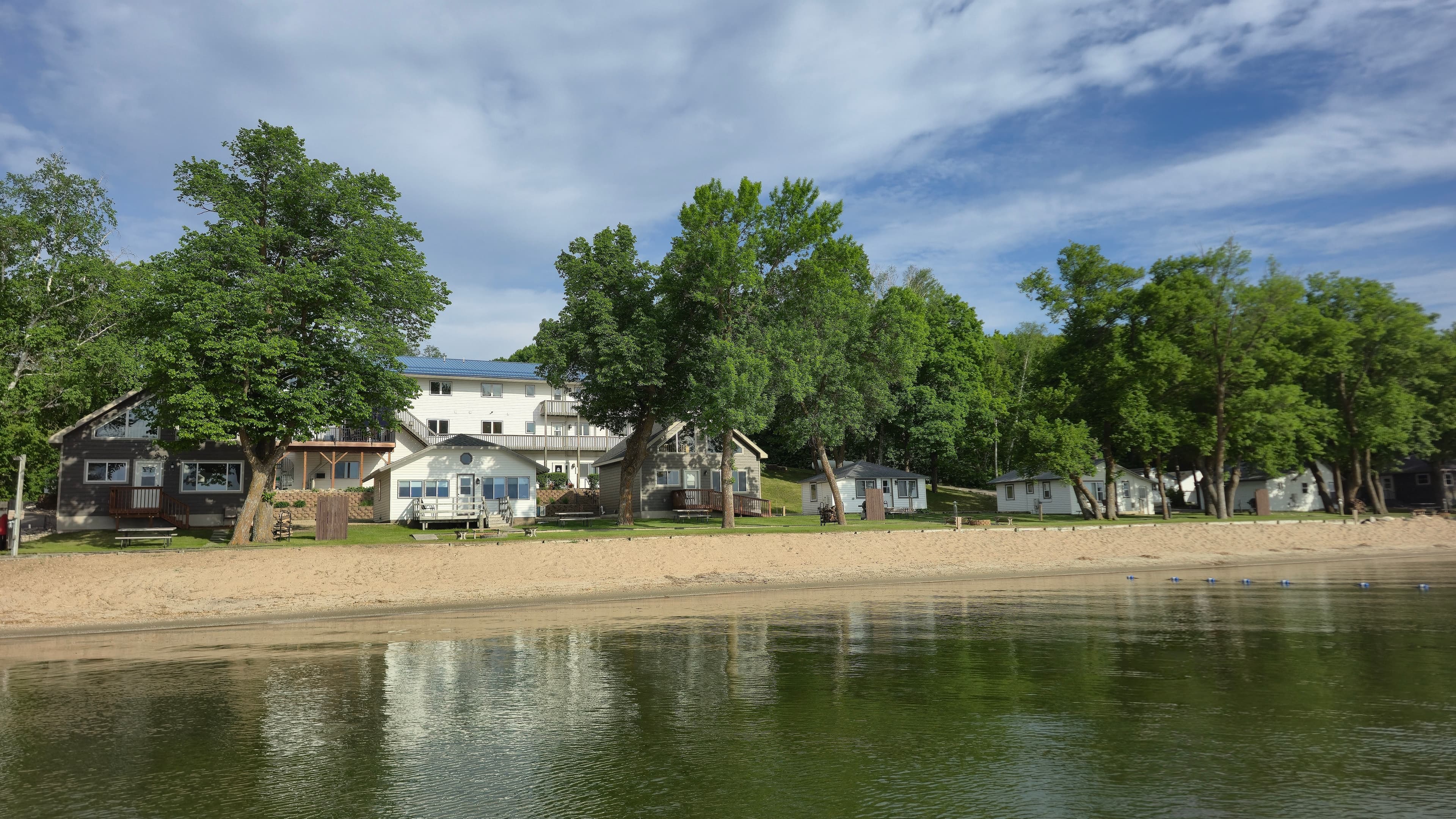 A row of houses bordered by a sandy beach and lush trees alongside a calm lake under a partly cloudy sky.